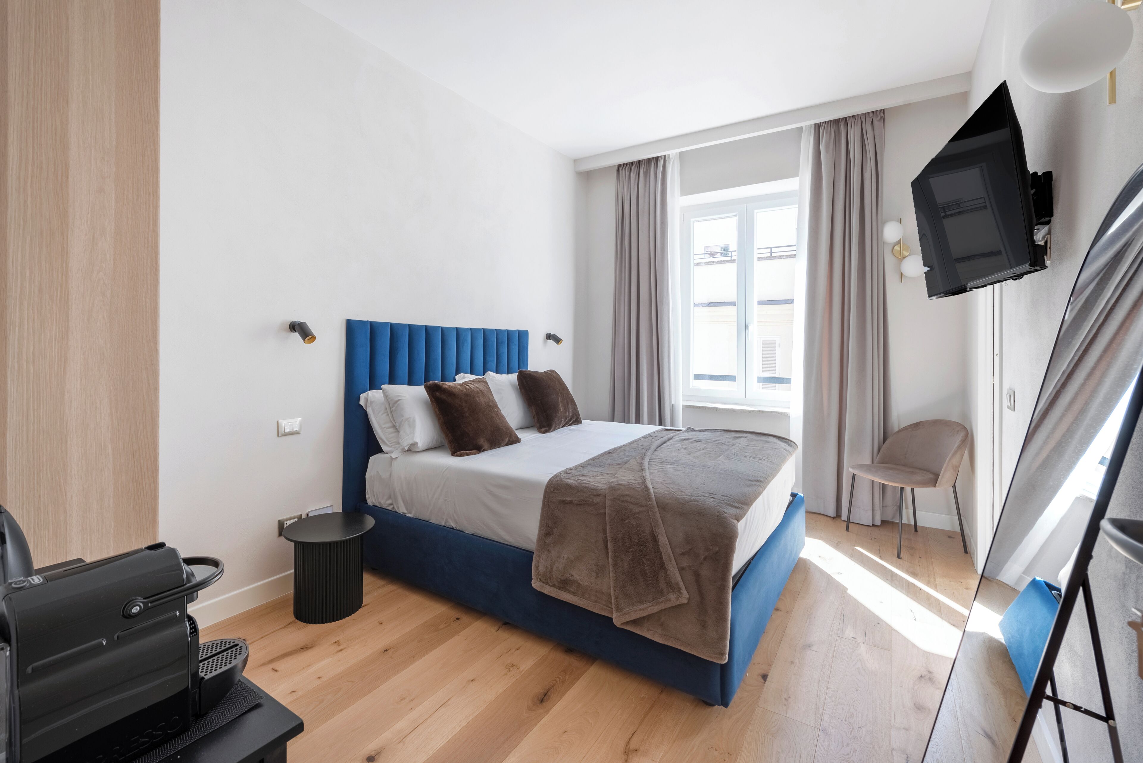 Une chambre d’hôtel moderne avec un lit bleu, un téléviseur fixé au mur, une petite table et une chaise.