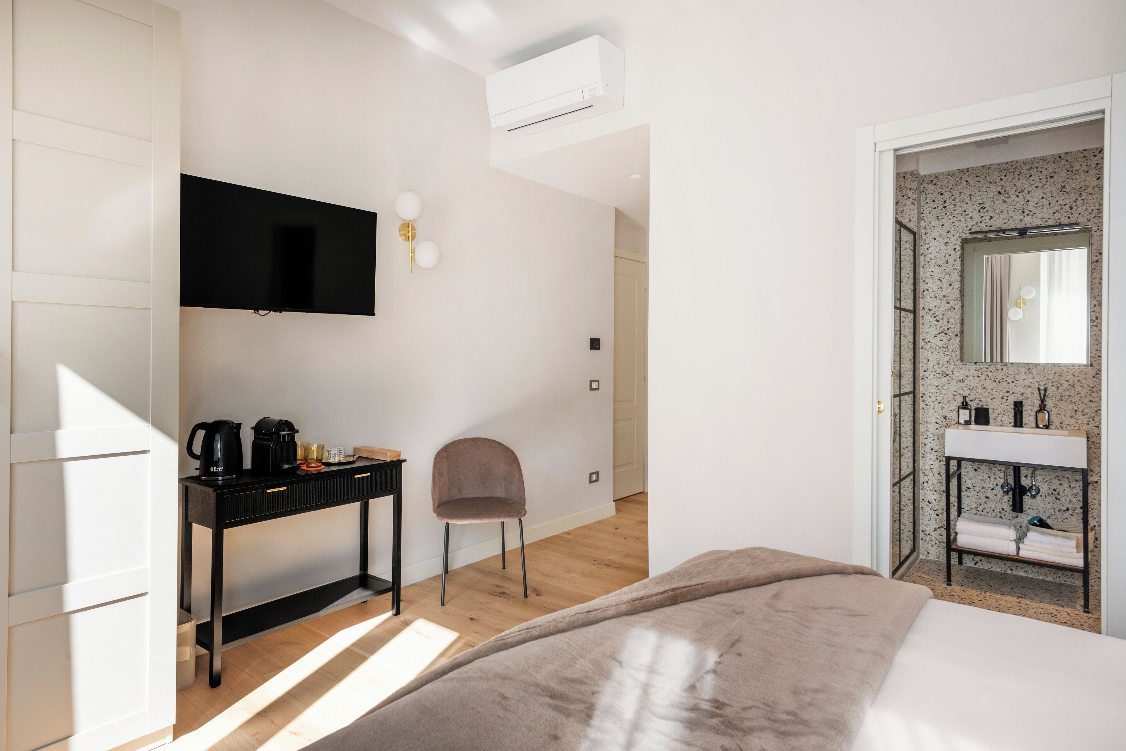 Een moderne hotelkamer met een bed, een bureau met koffiezetapparaat, een stoel, een aan de muur bevestigde televisie en een badkamer met wastafel en spiegel.