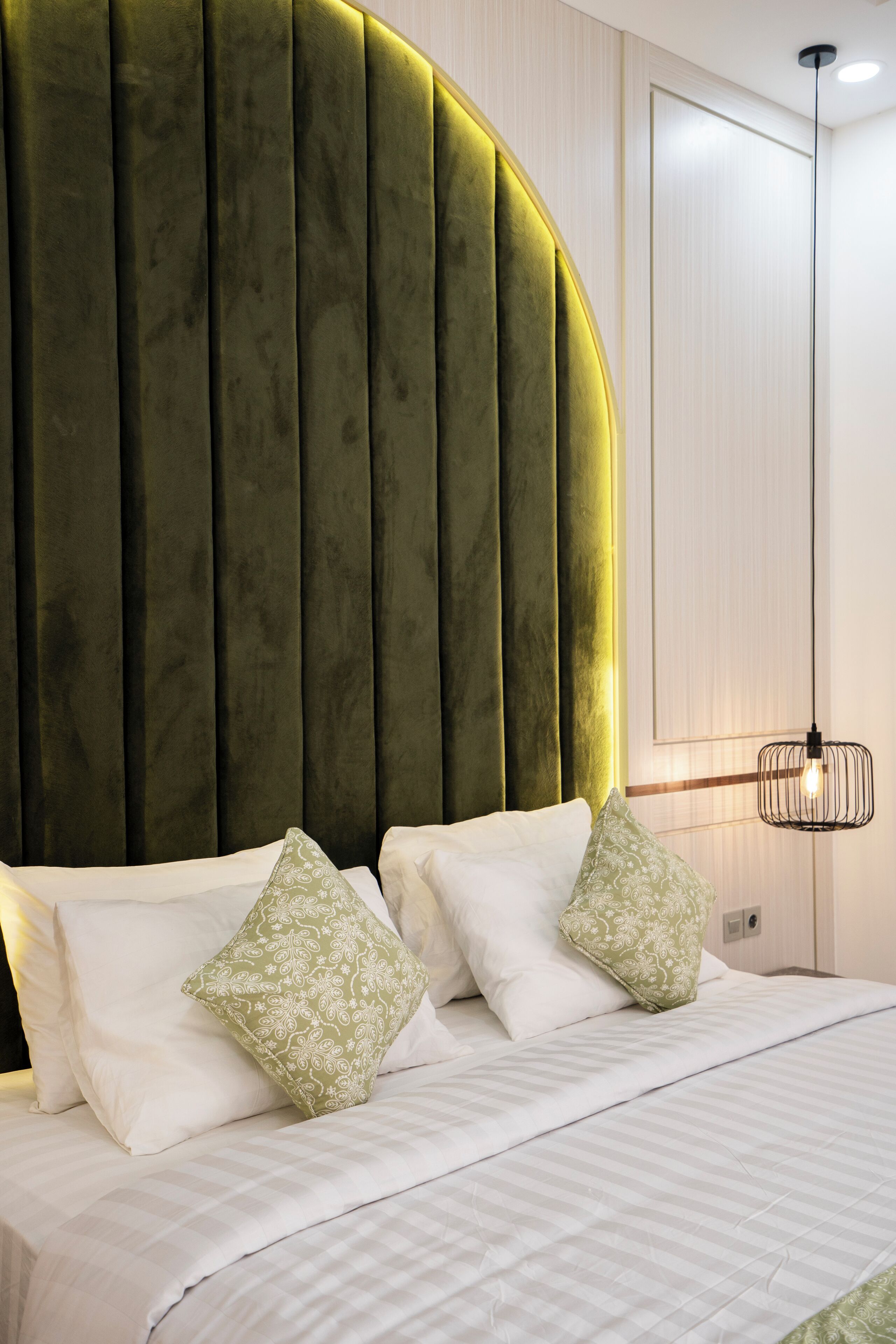 A bed with white linens and two patterned pillows, a green upholstered headboard, and a pendant light fixture.