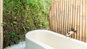 Una vasca da bagno sotto un tetto di paglia, circondata da pareti di bambù e vegetazione.