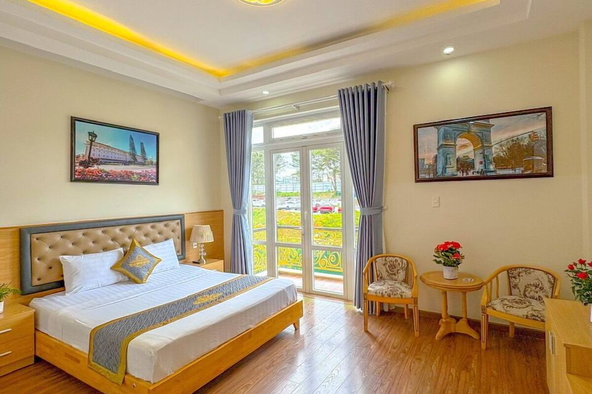 A hotel room with a large bed, a balcony with curtains, and two chairs.