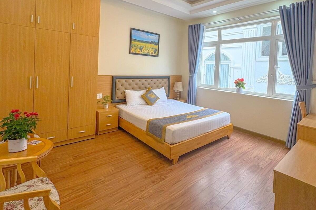 A bedroom with a large bed, wooden wardrobe, a small table with a plant, and a window with curtains.