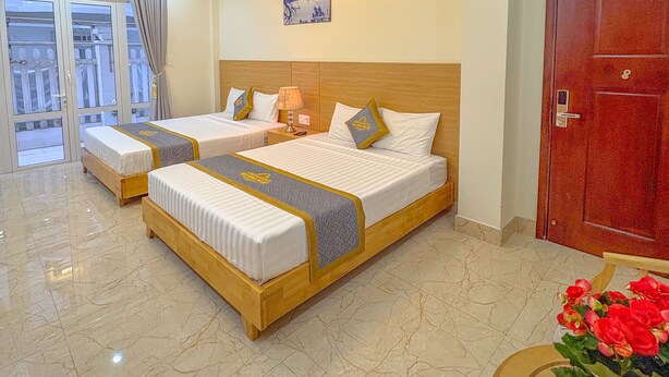 Ein Hotelzimmer mit zwei Betten, einem Holz-Kopfteil, einem großen Fenster mit Vorhängen, einer roten Tür und einem kleinen Tisch mit Blumen.