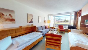 Une chambre d’hôtel comprenant un lit, un canapé, une chaise, une télévision et une fenêtre donnant sur des immeubles et des montagnes.