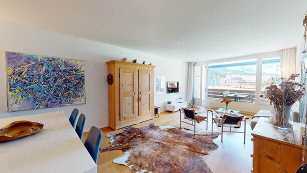Un salon moderne avec un grand tableau abstrait, un meuble en bois, un coin repas agrémenté d’un tapis en peau de vache, et une vue sur la ville grâce à de grandes fenêtres.