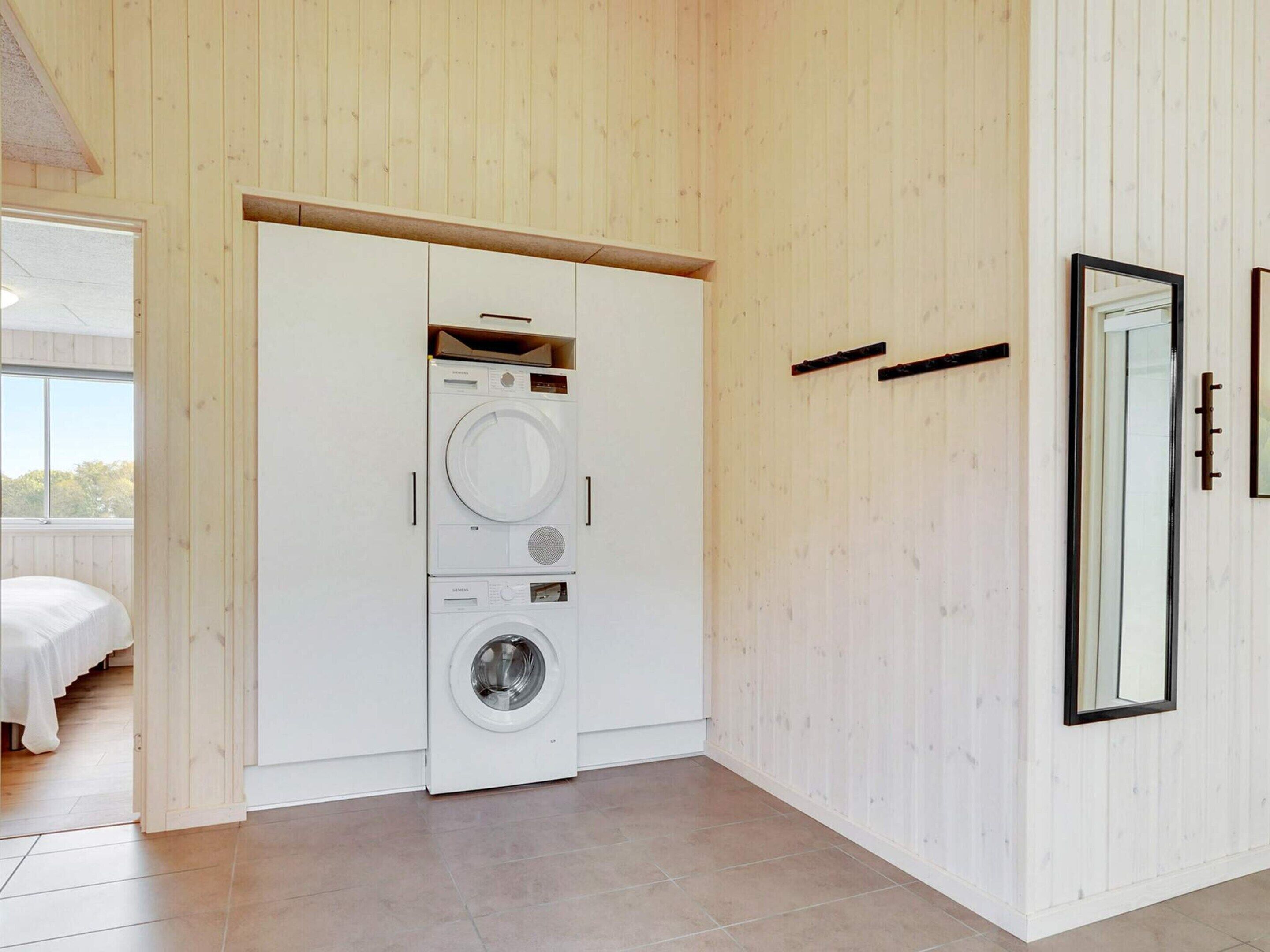 A laundry room with stacked washers and dryers, a bed in the adjacent room, and a large mirror on the wall.