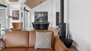 Un salon moderne avec un canapé en cuir, une table à manger avec des chaises, un poêle à bois noir et une grande fenêtre.