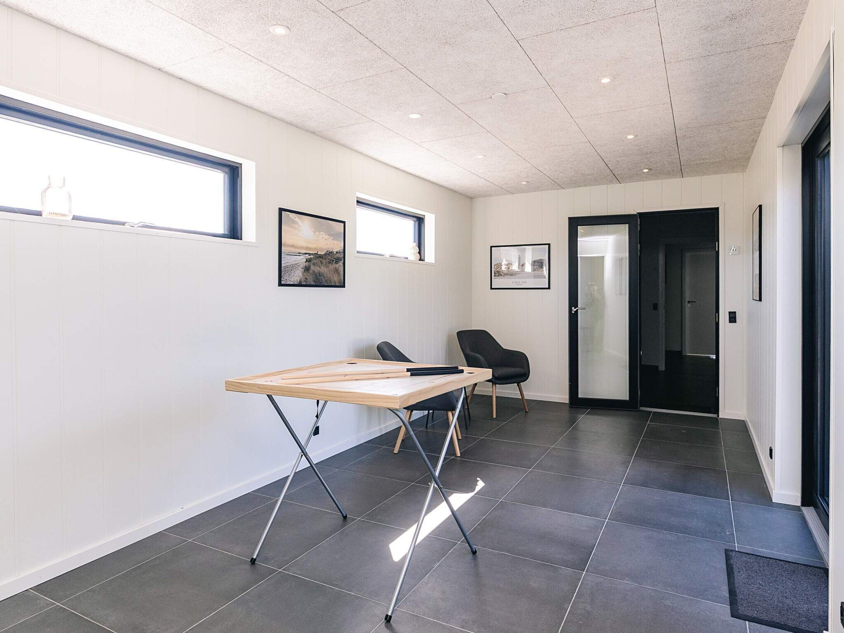 Ein moderner Arbeitsraum mit Stehschreibtisch, zwei Stühlen, gerahmten Bildern an der Wand und einer Tür zu einem anderen Zimmer.