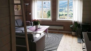 Et hyggeligt rum med træloft, et bord med en vase blomster, en sofa og to vinduer med udsigt over landskabet.