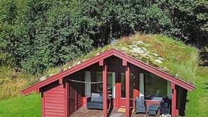 Une cabane en bois rouge, dotée d’un toit en pente, de grandes fenêtres et d’un petit espace porche aménagé avec des sièges d’extérieur.