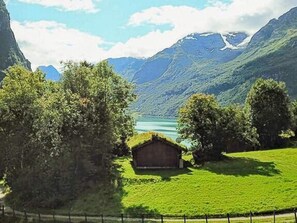 A rural landscape with a small wooden cabin surrounded by lush green fields and trees, overlooking a calm lake with mountains in the background. - 6 Person Holiday Home in Olden (Stryn)