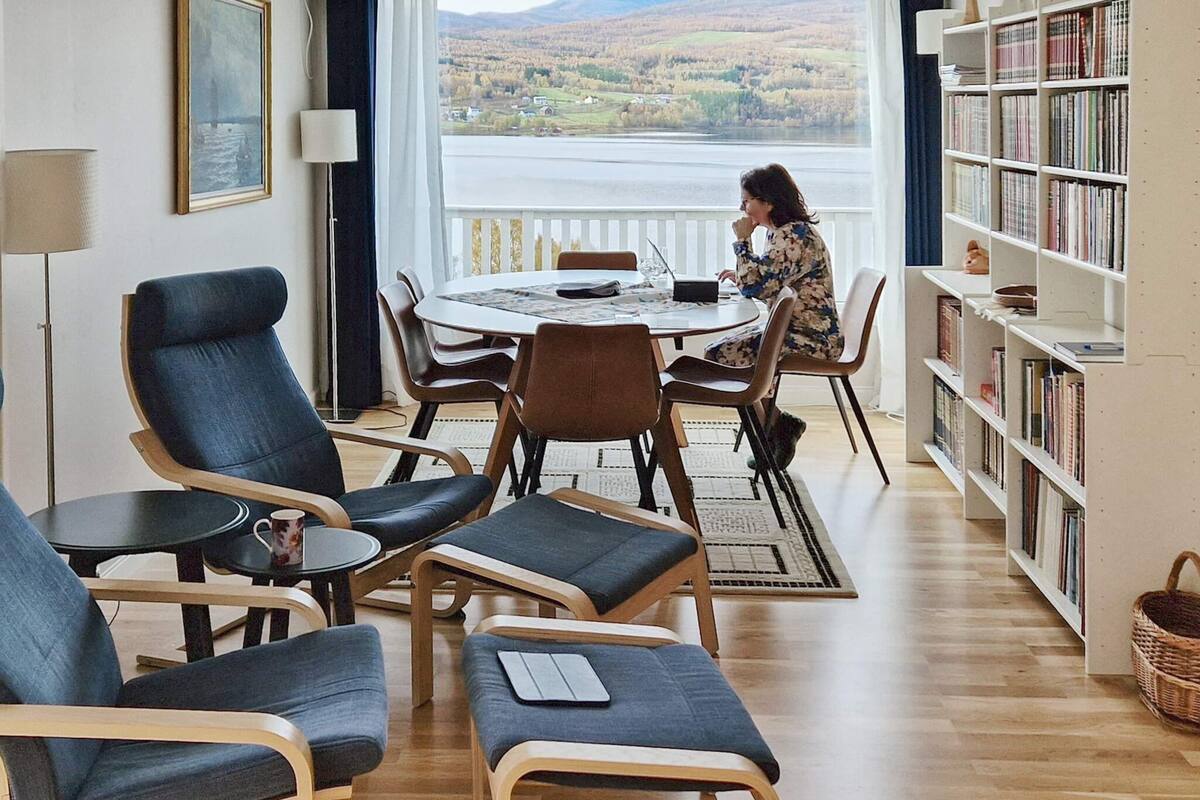 A modern living room with a view of a lake, a dining area with a round table, and a bookshelf filled with books.