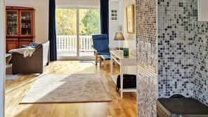 Un salon spacieux doté d’un plancher en bois, d’un canapé, d’une table basse et d’une cheminée. On y trouve également un coin repas avec une table et des chaises, et une vue sur l’extérieur grâce à de grandes fenêtres.