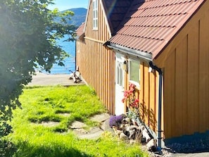 A wooden house with a sloped roof, a small porch, and a garden with flowers and shrubs. - 6 Person Holiday Home in Maloy (Kinn)