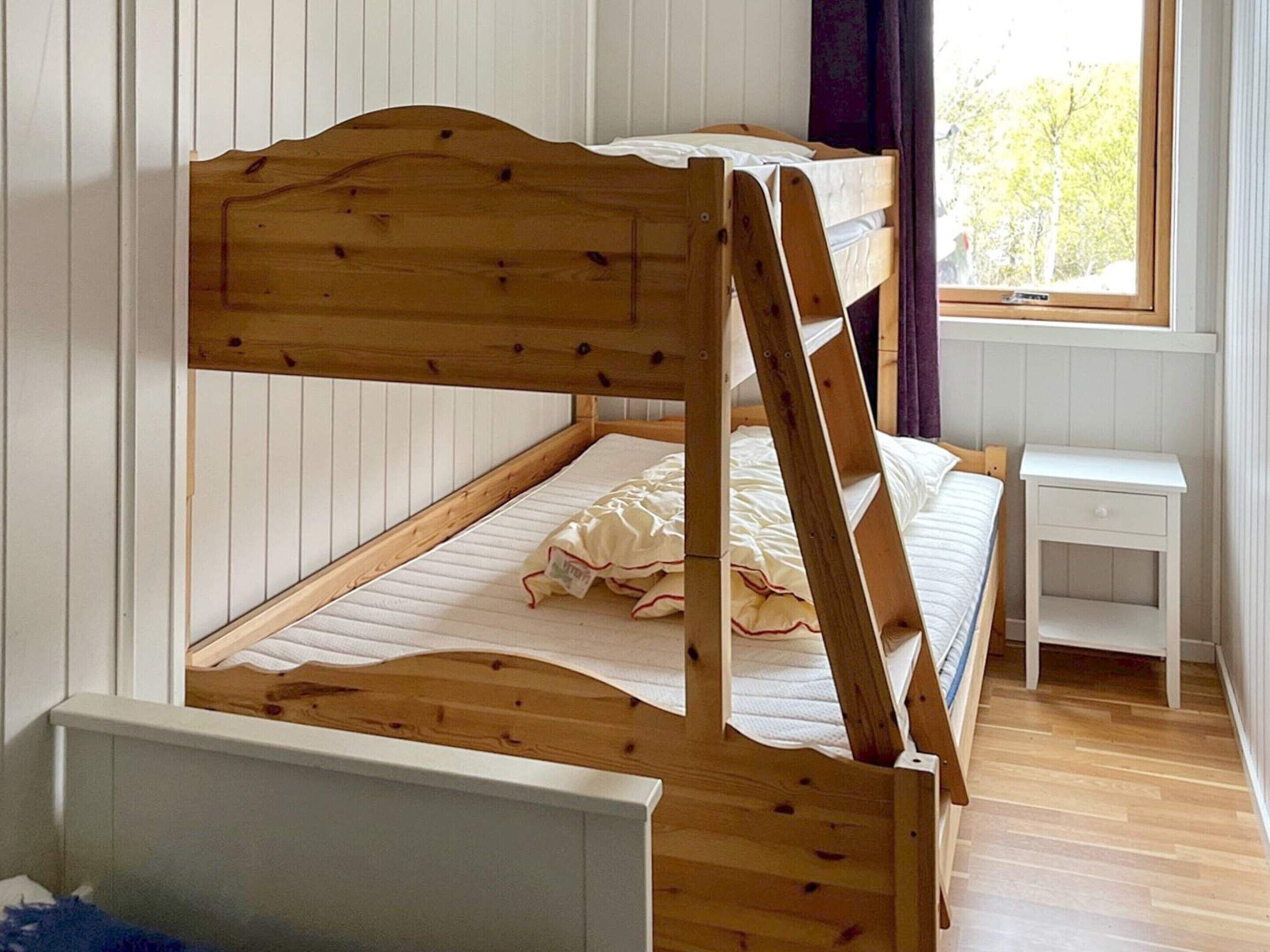 A bunk bed with a wooden frame, a bedside table, and a window with a view of trees.