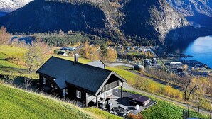 Une maison avec un toit sombre et une terrasse, entourée d’un paysage verdoyant luxuriant et dominée par un décor montagneux.