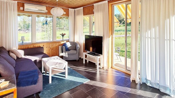 A cozy living room with a sofa, armchair, and TV, large windows, and a view of greenery.