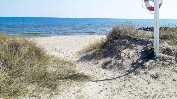 A beach with a lifebuoy attached to a pole, clear blue water, and sandy dunes.