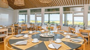 A dining area with round tables set for a meal, large windows offering a view of the beach, and a ceiling with woven light fixtures.