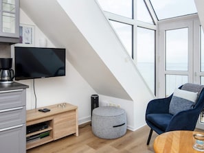 A modern loft bedroom with a large skylight, a television, a coffee maker, a small ottoman, and a blue armchair.