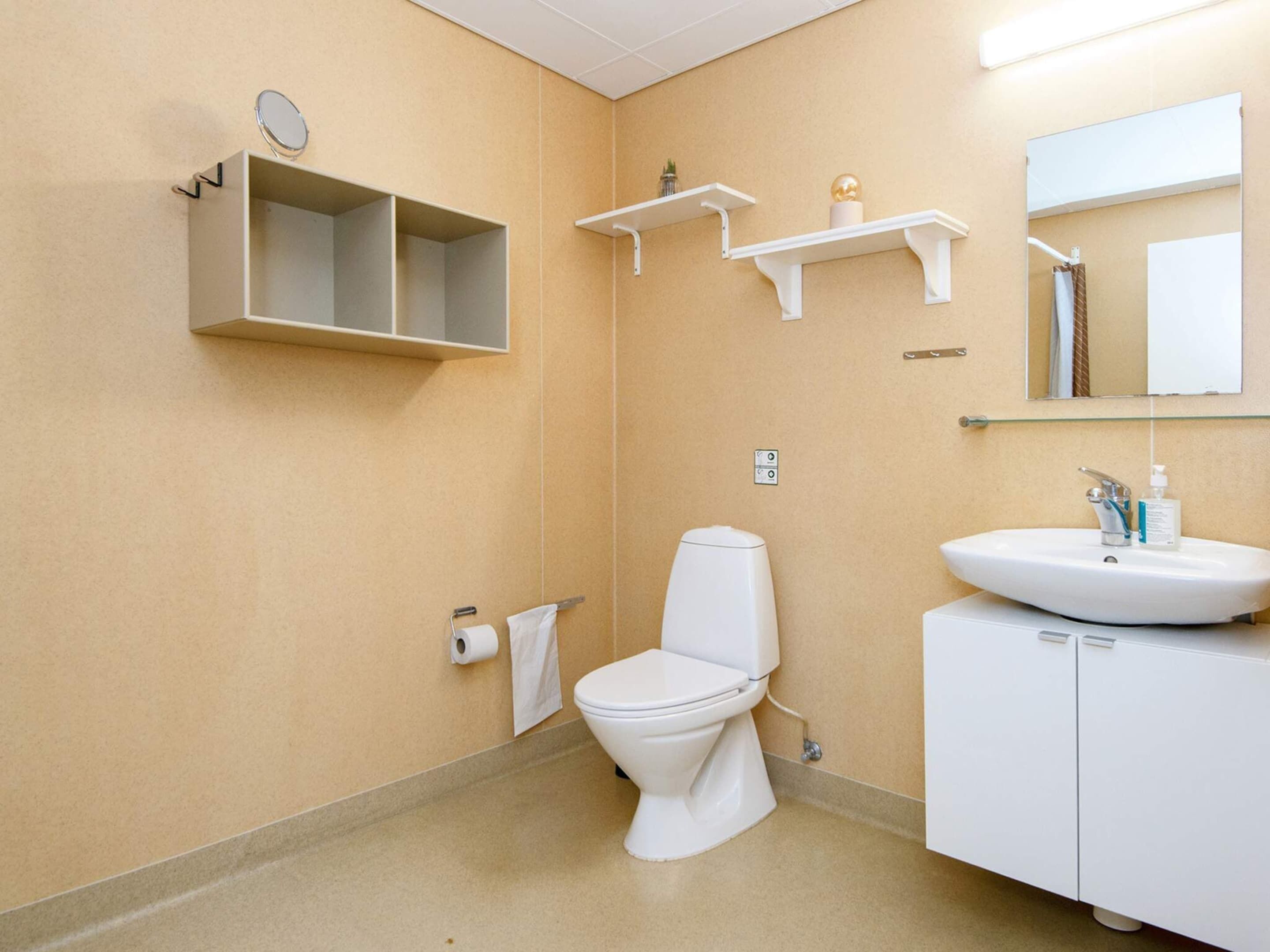 Une salle de bain avec des toilettes, un lavabo et un miroir. Il y a des étagères au-dessus des toilettes et un petit miroir sur le mur.