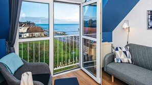 Habitación con un amplio ventanal corredero que da a un balcón con vistas al mar y edificios. Se aprecia un sofá gris y una silla con cojín.