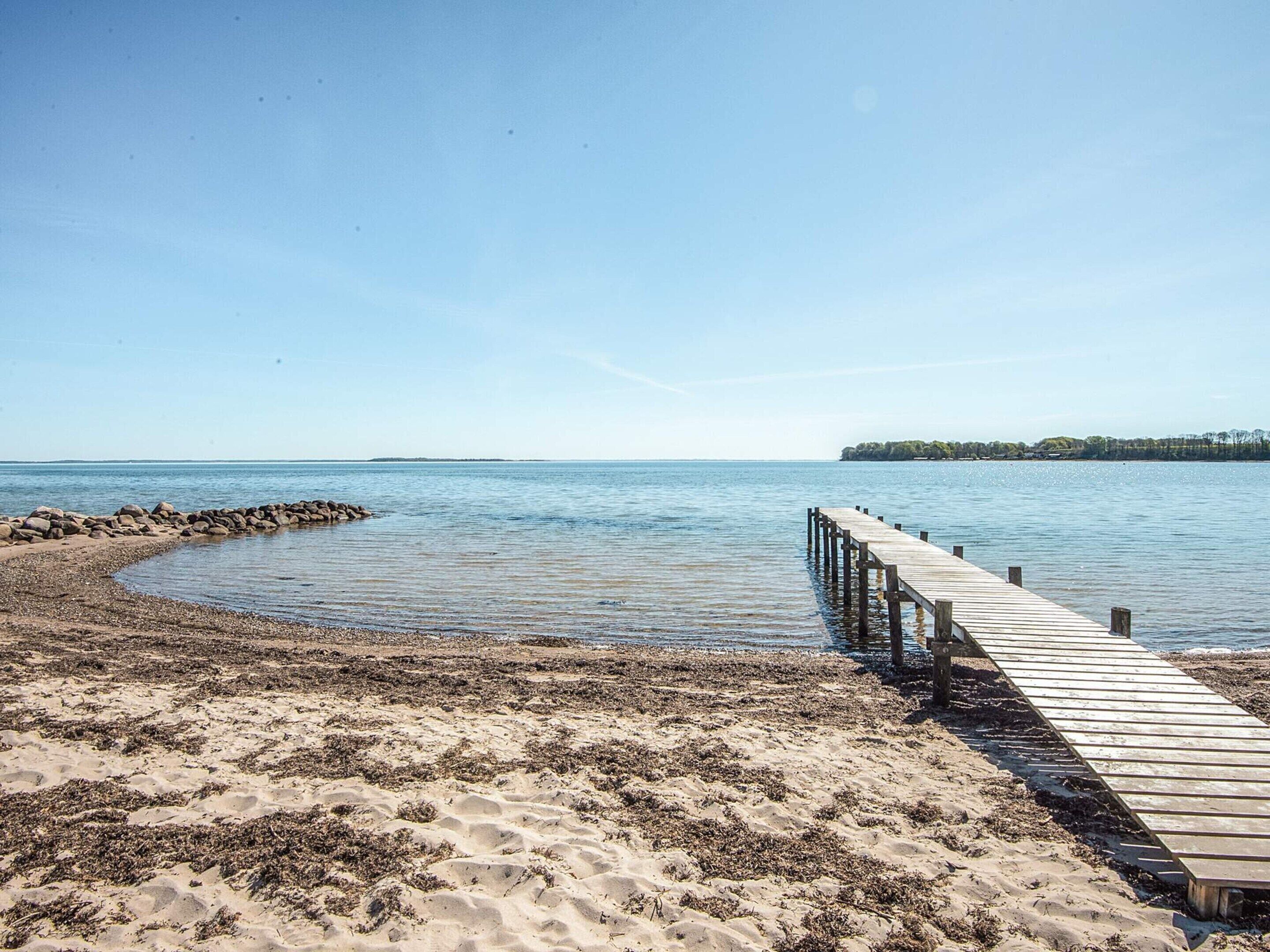 Un muelle de madera se adentra en aguas tranquilas, rodeado de una playa arenosa con algas.
