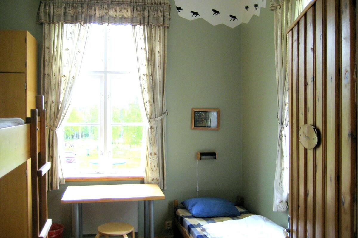 Ett litet rum med en enkel säng, ett fönster, ett skrivbord och en stol.