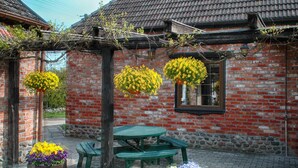 A rustic brick building with a tiled roof, a wooden table, and benches. There are flower pots with yellow and purple flowers. - Stylish House in the Countryside 5 km From the sea (Darlowo)