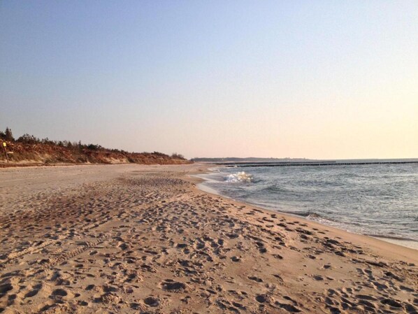 A sandy beach with footprints leading to the water, a clear sky, and a distant shoreline with vegetation. - Stylish House in the Countryside 5 km From the sea (Darlowo)