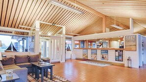 Eine großzügige Lobby mit hoher Holzbalkendecke, einer Rezeption, einer Sitzecke mit Sofa und Tisch sowie Blick durch große Fenster.