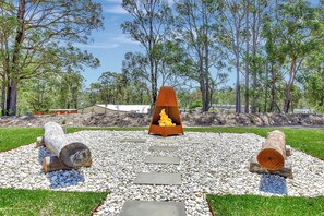 A fire pit surrounded by logs and stones, set in a landscaped area with trees and a clear sky.