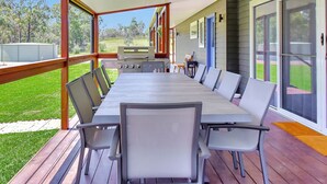 Une terrasse extérieure couverte, dotée d’une longue table à manger et de chaises, d’un barbecue et offrant une vue sur une pelouse.