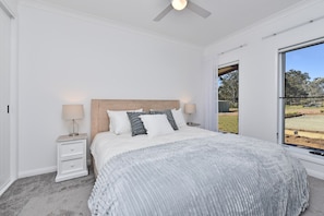 A bedroom with a bed, nightstands, a ceiling fan, and a large window with a view of the outdoors.