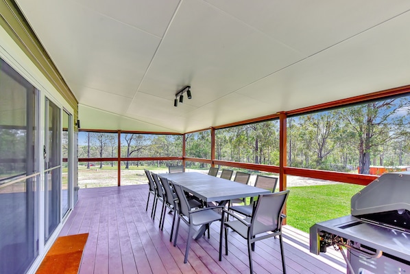 A covered outdoor patio with a dining table and chairs, a grill, and a view of a grassy area with trees.