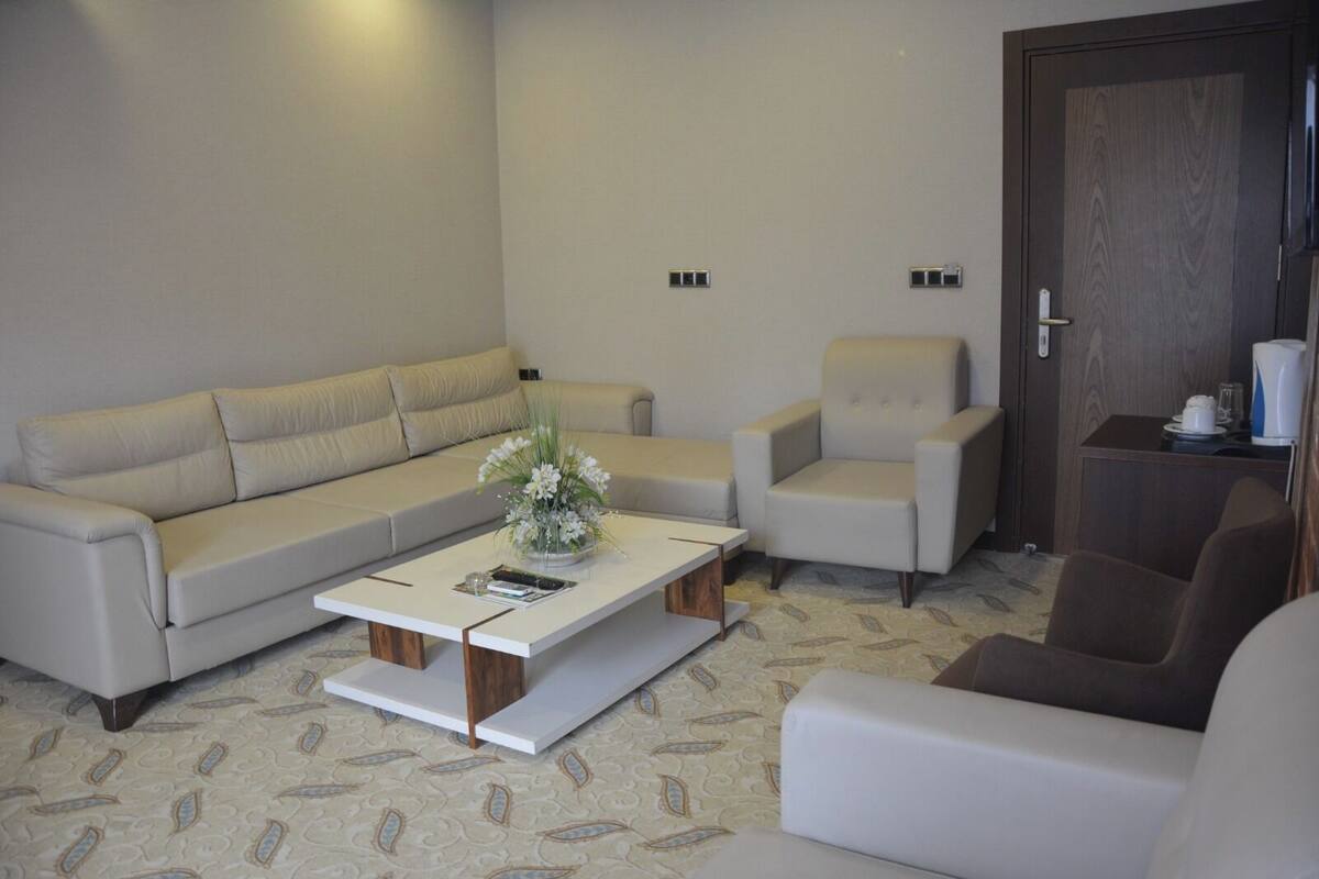 A living room with a sofa, two armchairs, a coffee table with a vase of flowers, a door, and a small table with a kettle and cups.