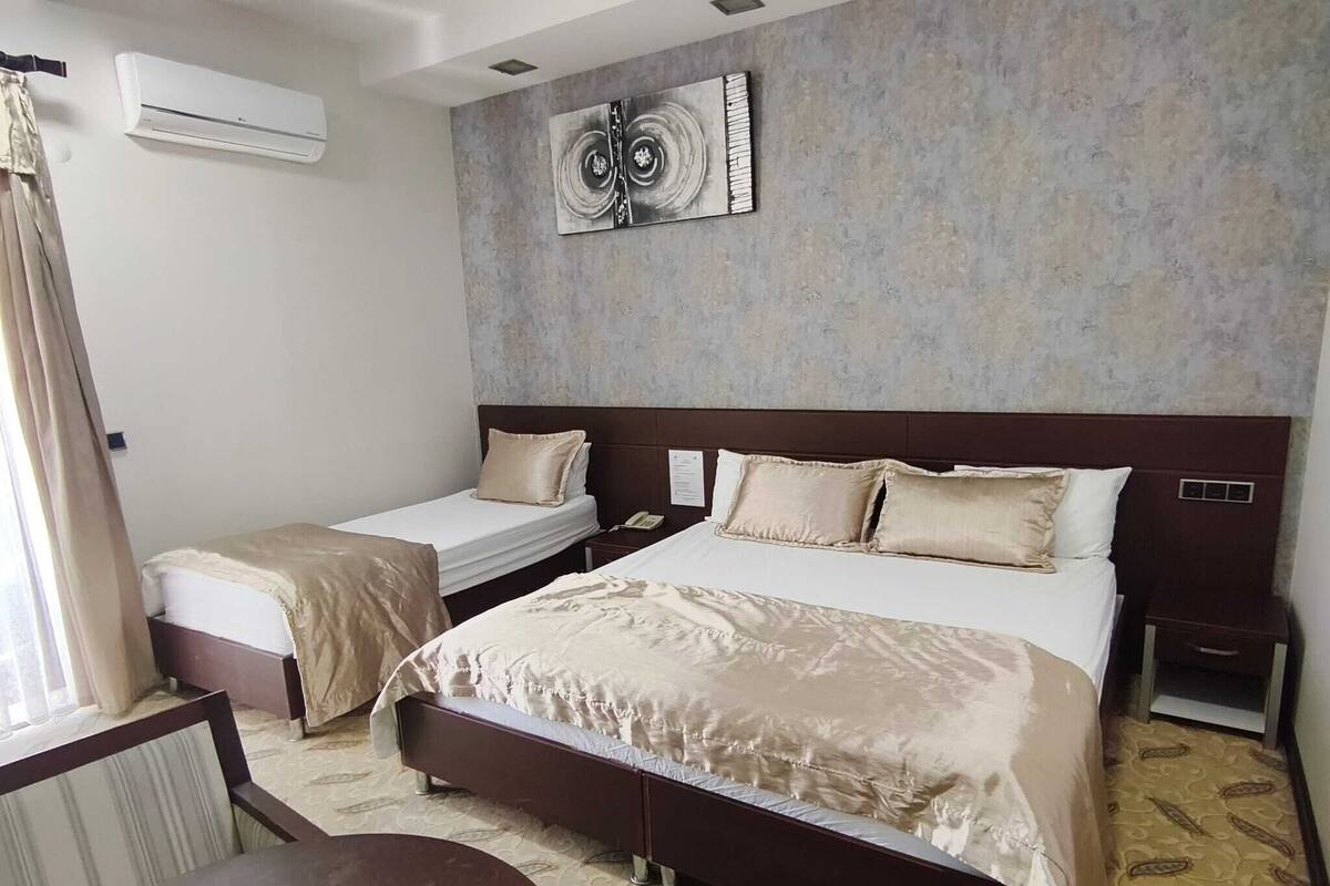 A hotel room with a large bed, a smaller bed, a bedside table, a chair, a desk, and an air conditioning unit.