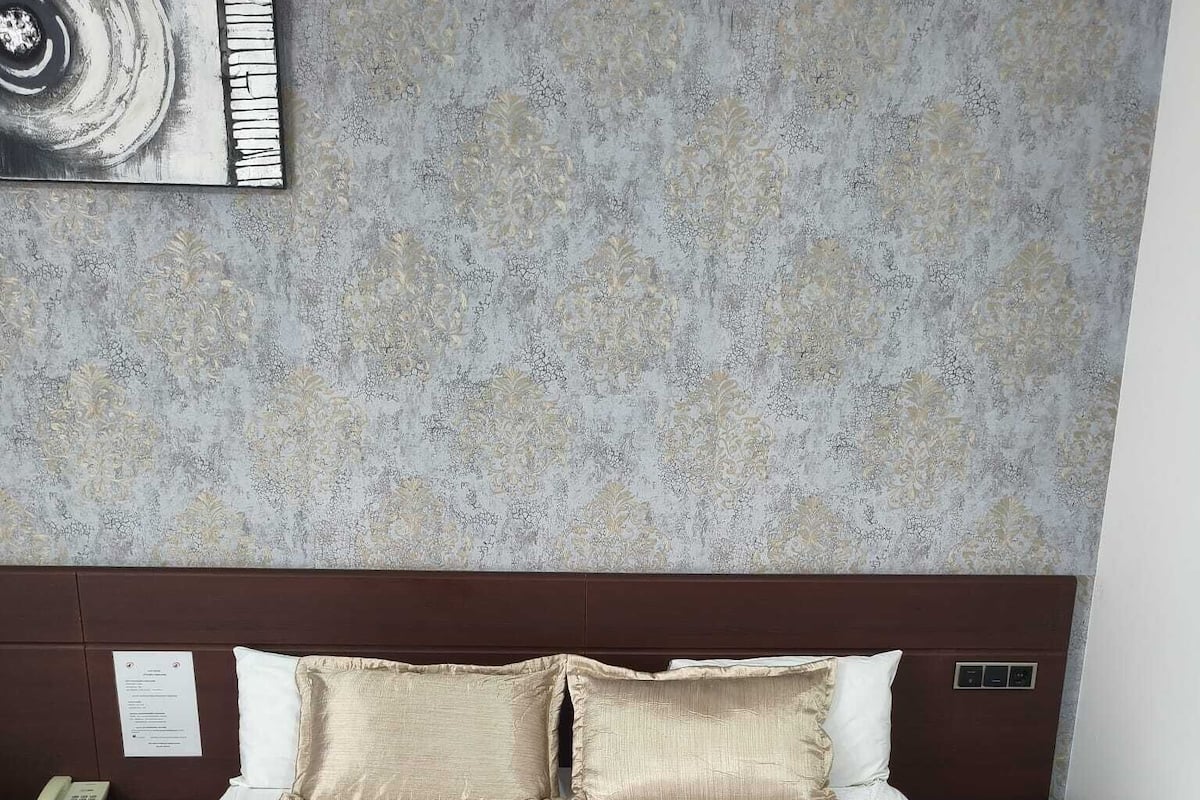 A neatly made bed with a beige bedspread and two pillows, a wooden headboard, a bedside table with a lamp, and a wall-mounted artwork.