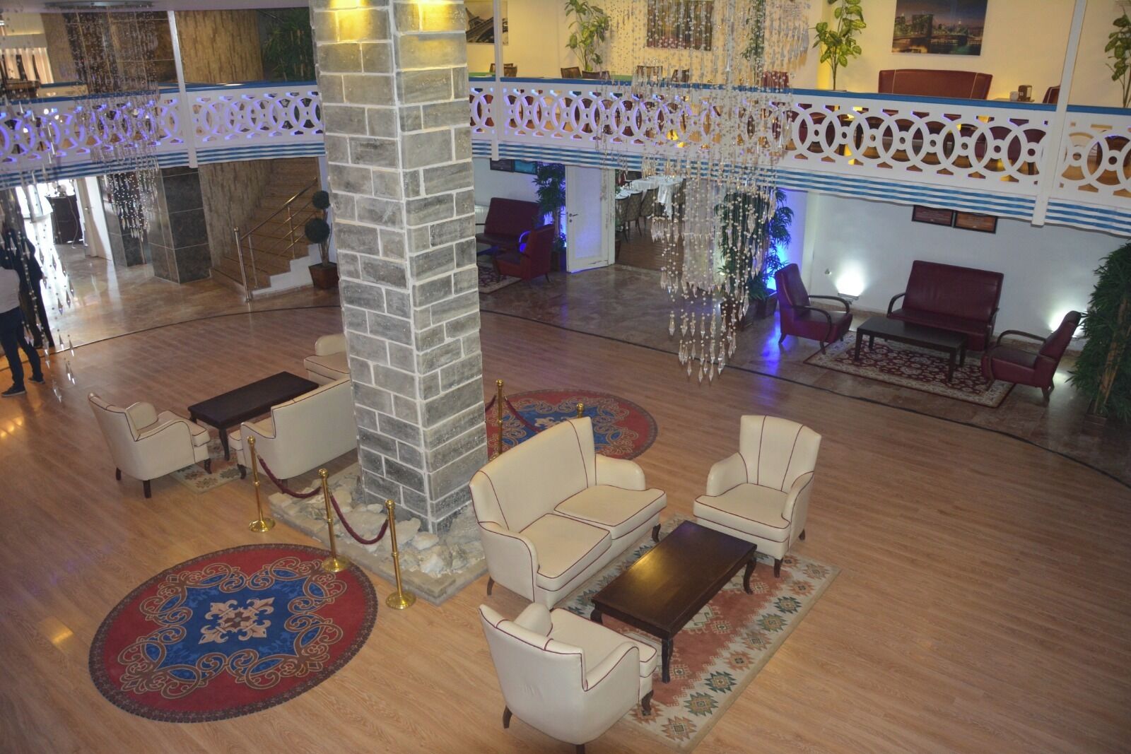 A spacious lobby with a high ceiling, a central stone pillar, and a chandelier. There are multiple seating areas with sofas and armchairs, and a dining area in the background.