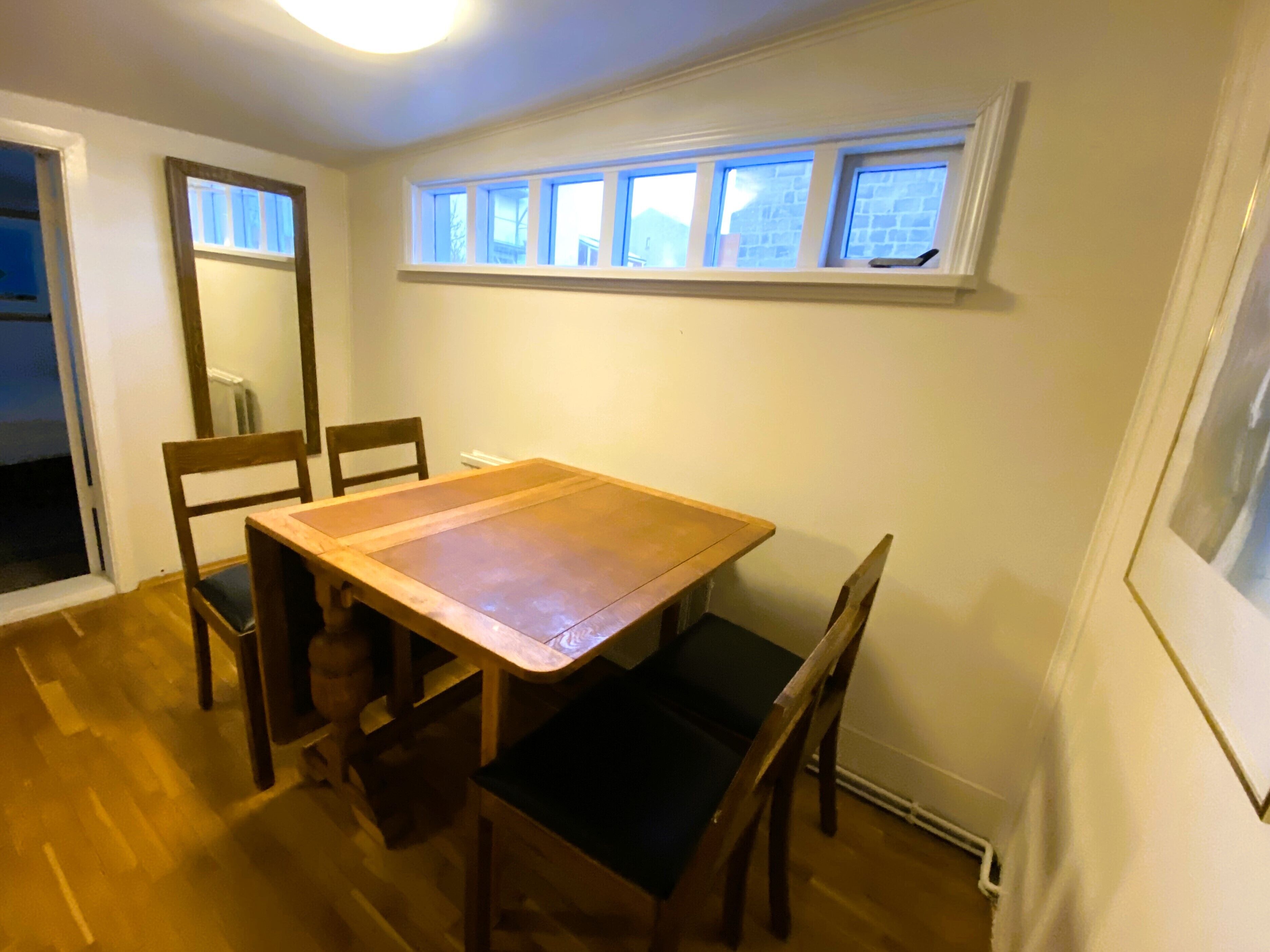 Un coin repas avec une table en bois et quatre chaises, une fenêtre donnant sur des immeubles et une porte menant à une autre pièce.