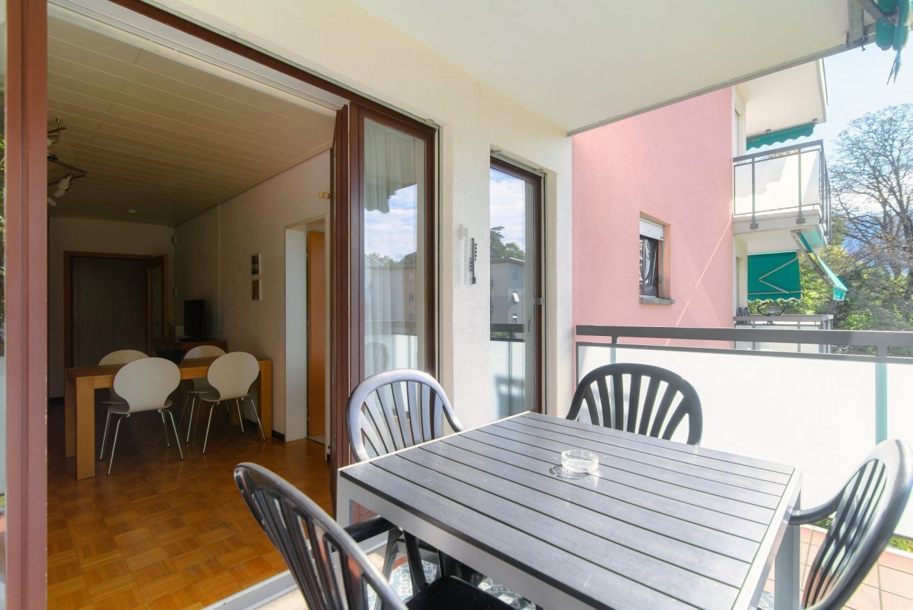 Un balcon avec une table et des chaises, une porte-fenêtre coulissante donnant sur un espace de repas intérieur, et un bâtiment rose en arrière-plan.