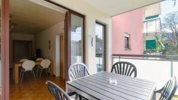 Un balcon avec une table et des chaises, une porte-fenêtre coulissante donnant sur un espace de repas intérieur, et un bâtiment rose en arrière-plan.