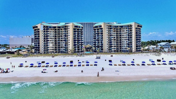 Un hôtel en bord de mer, avec plusieurs étages, une plage de sable dotée de parasols et une vue dégagée sur l’océan.