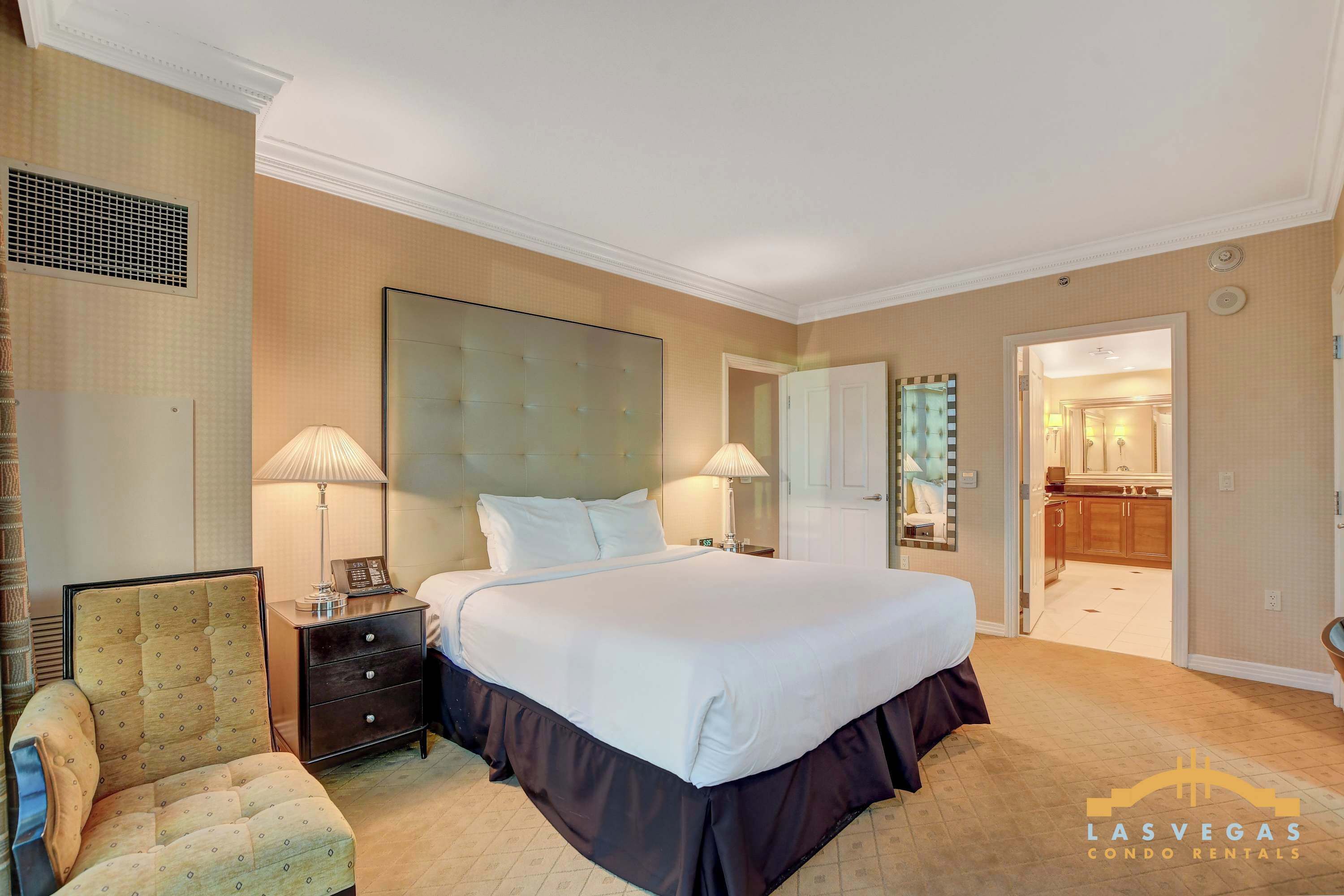 A hotel bedroom with a large bed, bedside lamps, a chair, and a bathroom visible through an open door.