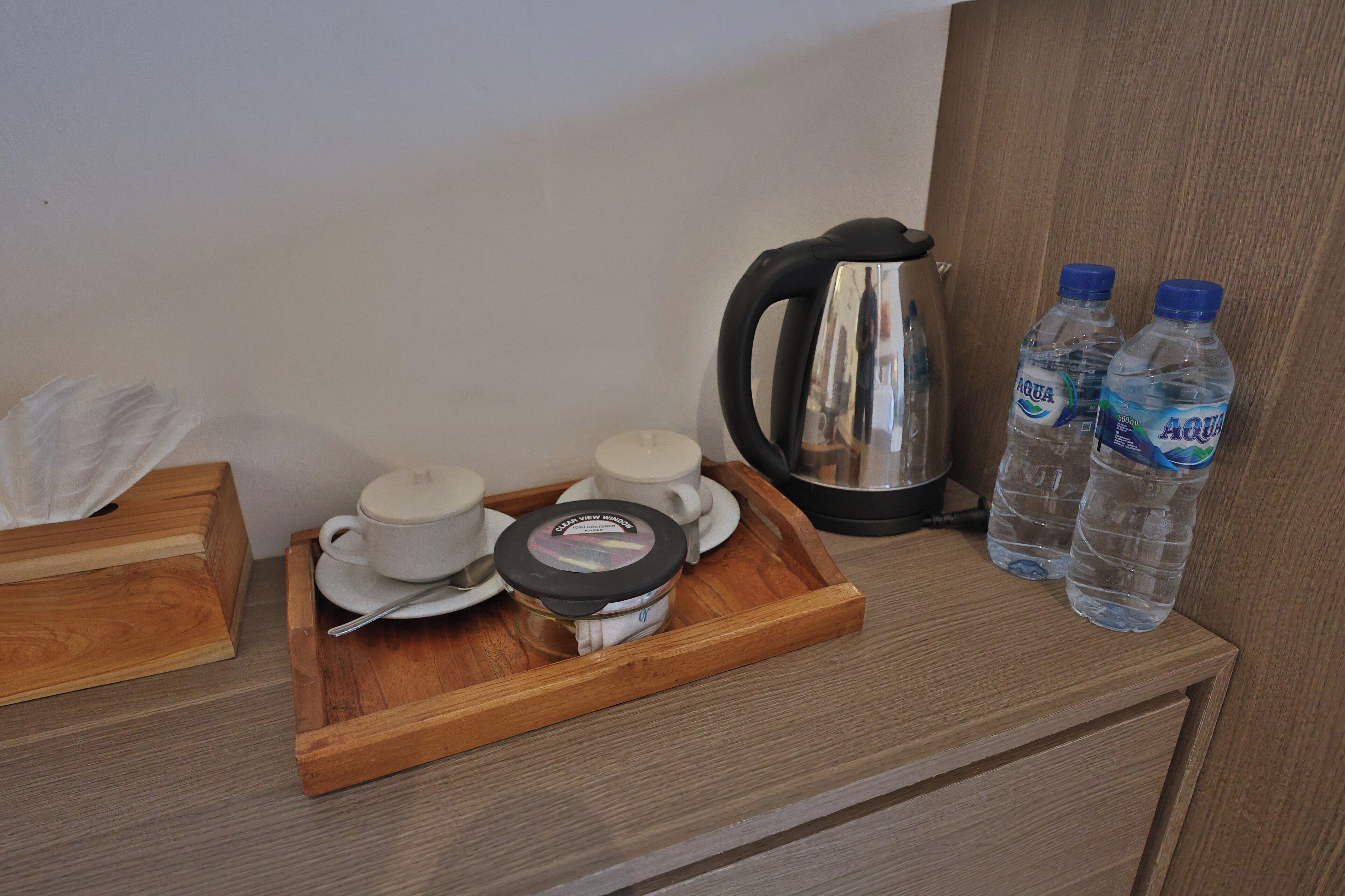 A wooden tray with a teapot, cups, and a jar of cream on a wooden table. Two bottles of water and a kettle are also visible.