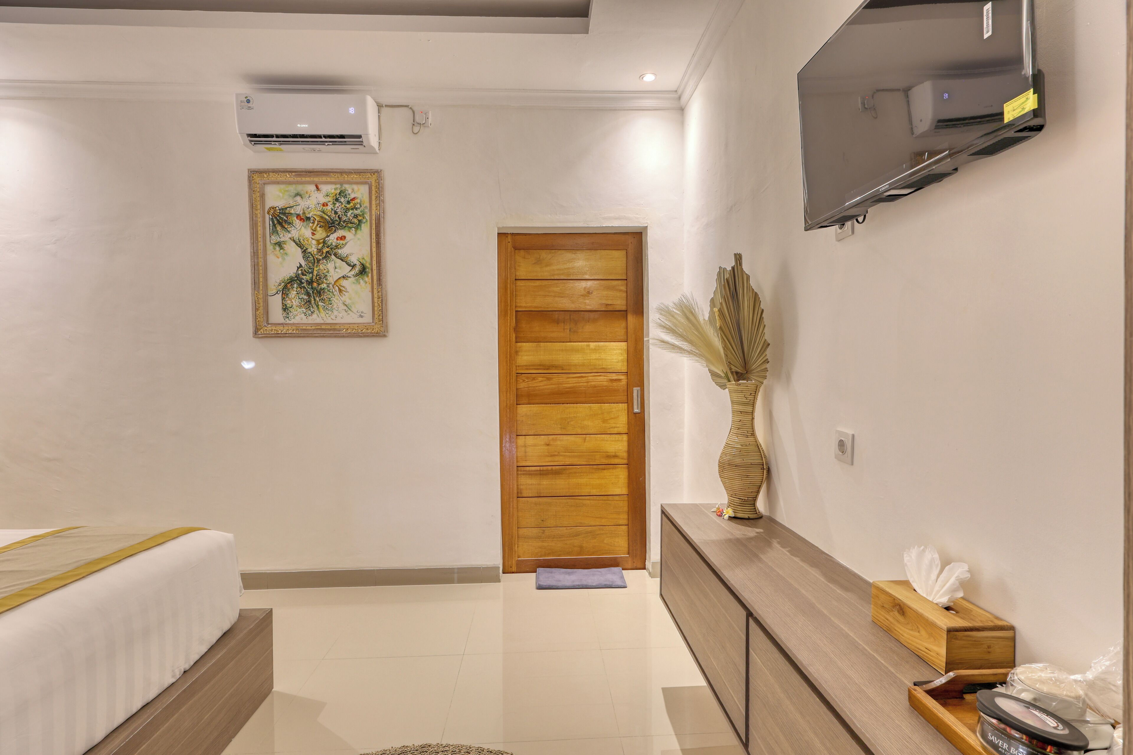 Una habitación de hotel moderna con una cama, una puerta de madera, un televisor montado y un jarrón con plantas secas.