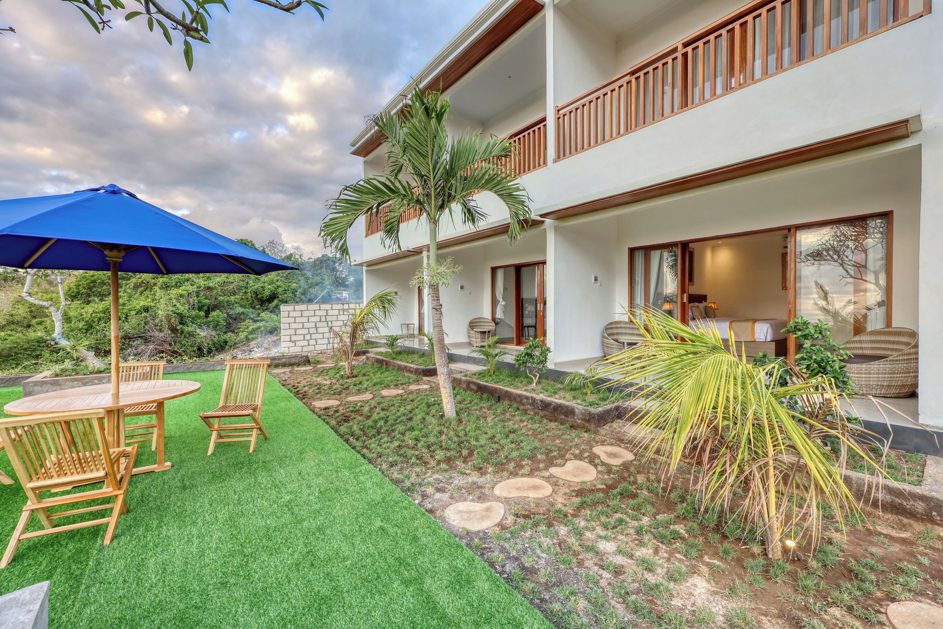 A two-story building with a balcony, a patio with artificial grass and wooden furniture, and a garden area with palm trees and stepping stones.
