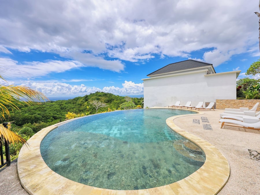 A clear swimming pool with a curved edge, surrounded by lounge chairs and a white building with a sloped roof.