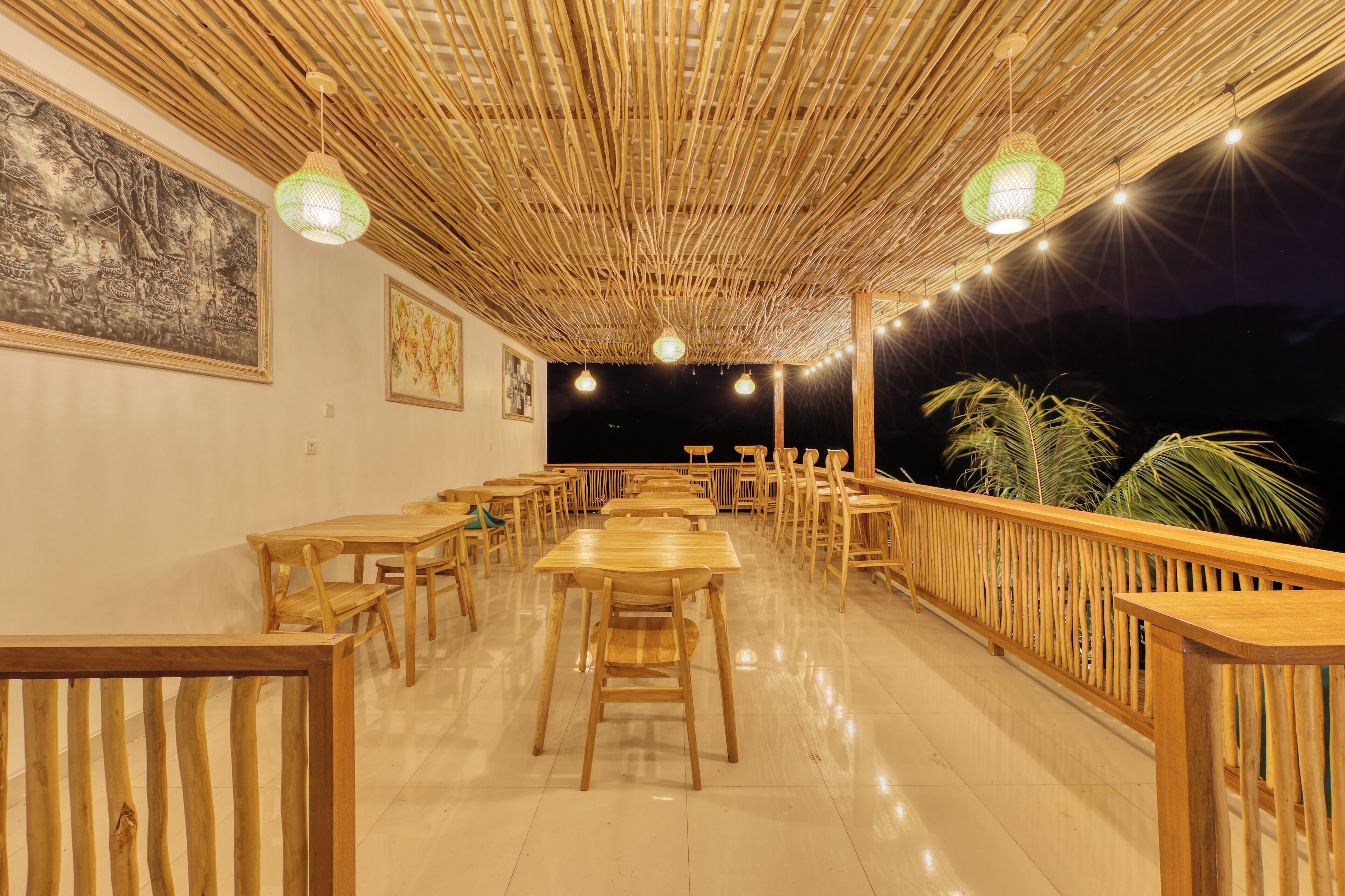 Un área de comedor interior con mesas y sillas de madera, techo de bambú y vistas a palmeras al exterior.