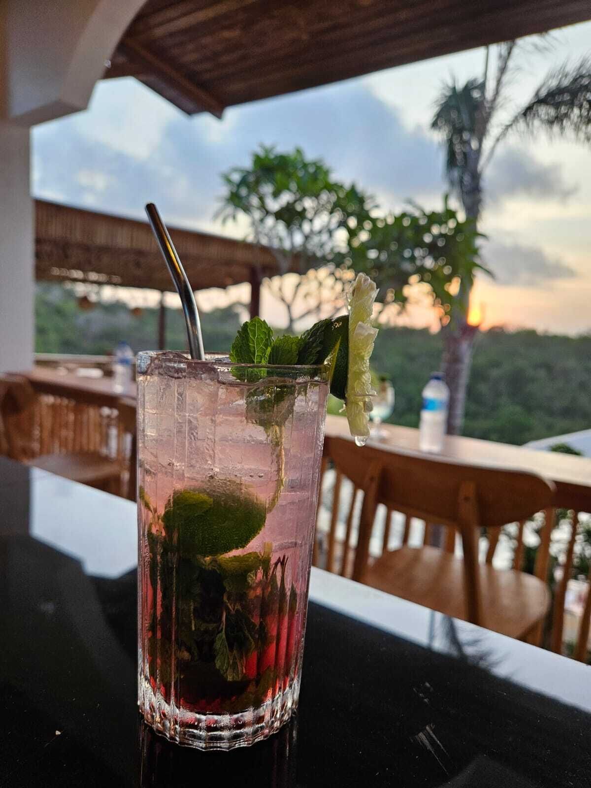 A glass of iced beverage with mint leaves and a lime wedge on a table with a view of trees and a sunset.